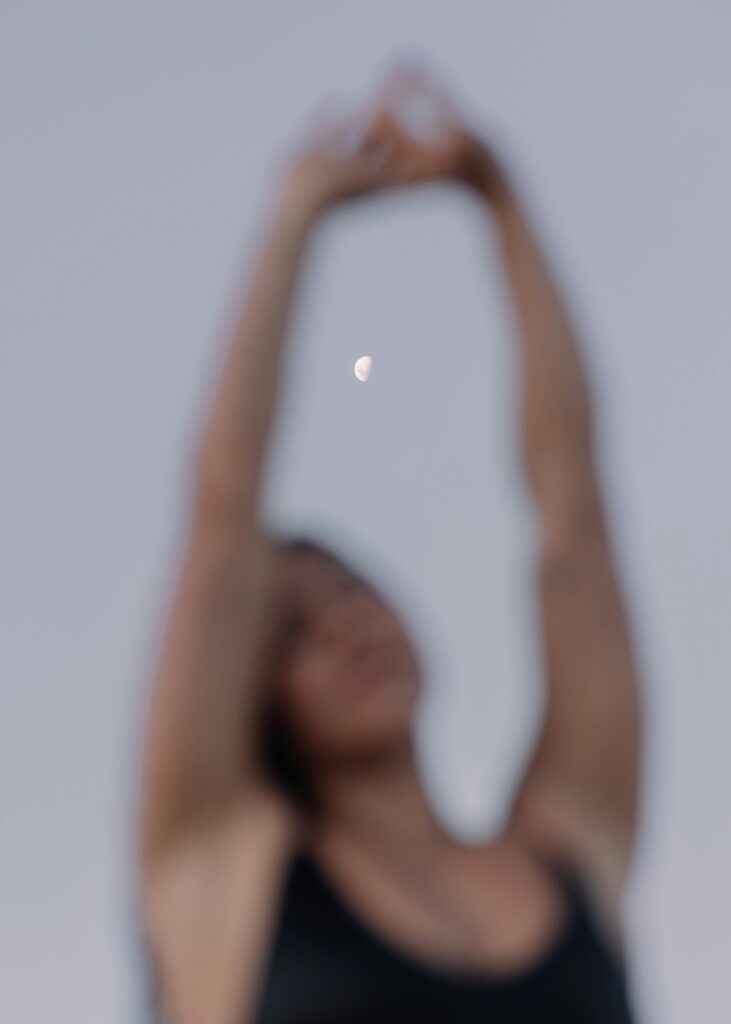A woman stretches with arms raised, silhouetted against a crescent moon and evening sky.