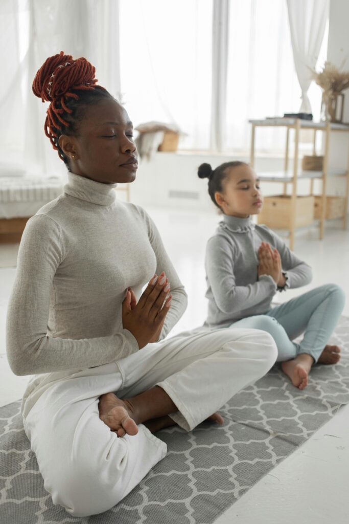pexels-photo-7353048-7353048 Full body of tranquil young barefooted African American woman in casual clothes sitting on carpet in Padmasana pose with closed eyes while meditating with adorable little daughter at home