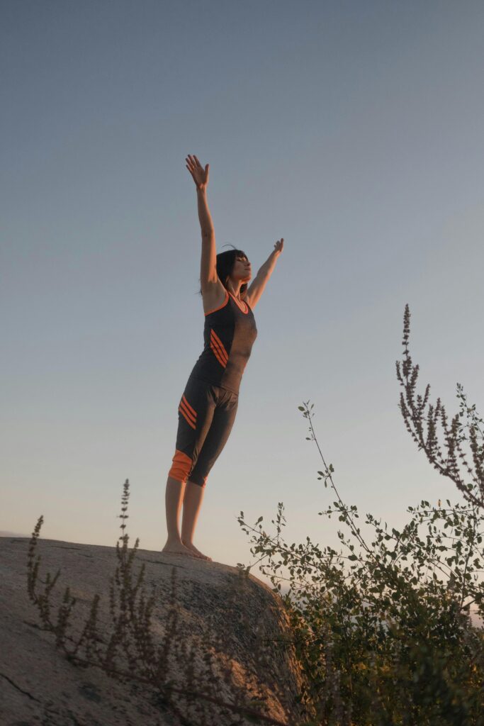 A woman in sportswear stands on a rock with arms raised, embracing nature at sunset.