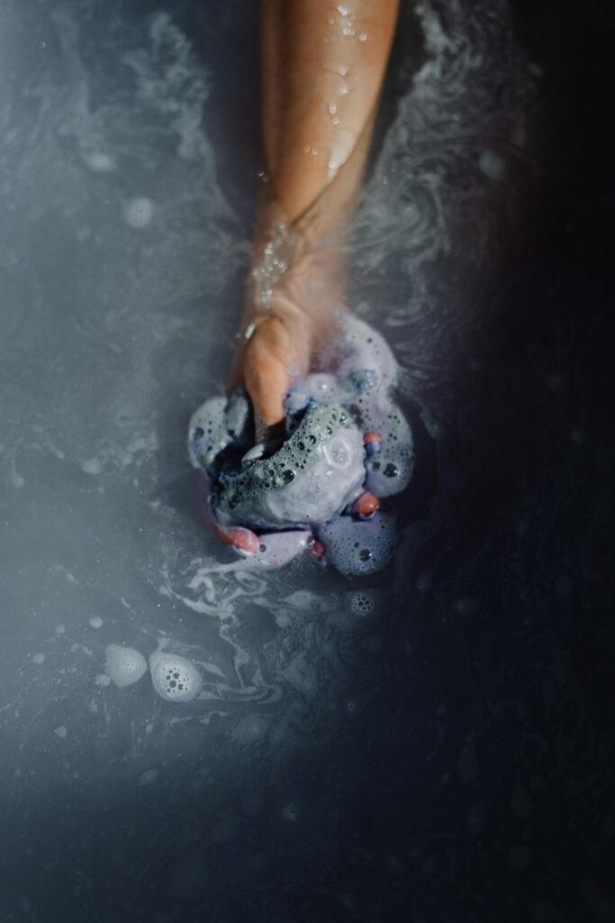 A hand holds a purple soap in a relaxing bath, surrounded by bubbles.